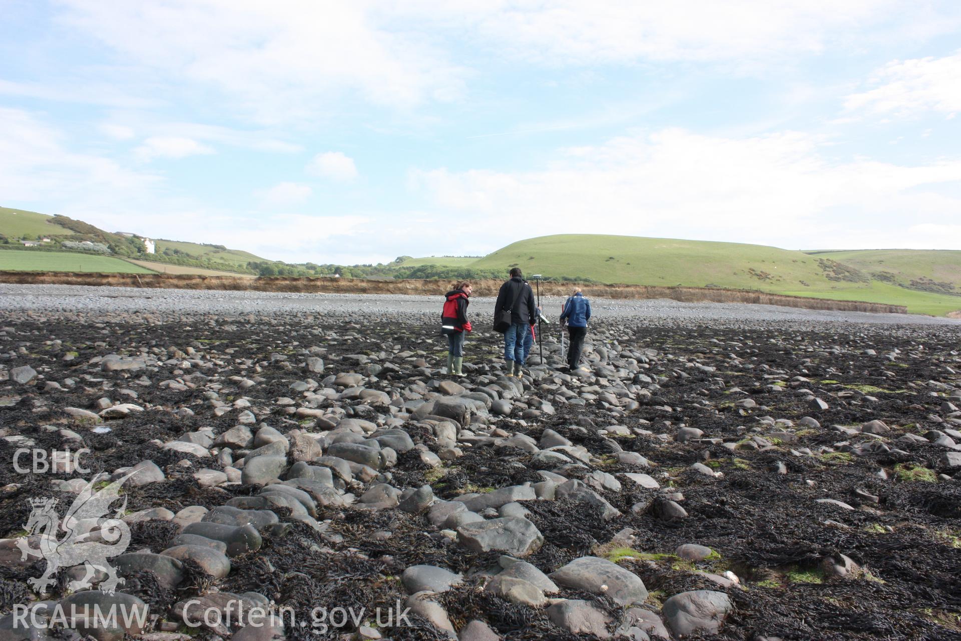 North-south arm of fish trap, looking south. Shows alignment of boulders comprising dry stone wall. Members of RCAHMW staff and local resident standing on alignment of fish trap arm.