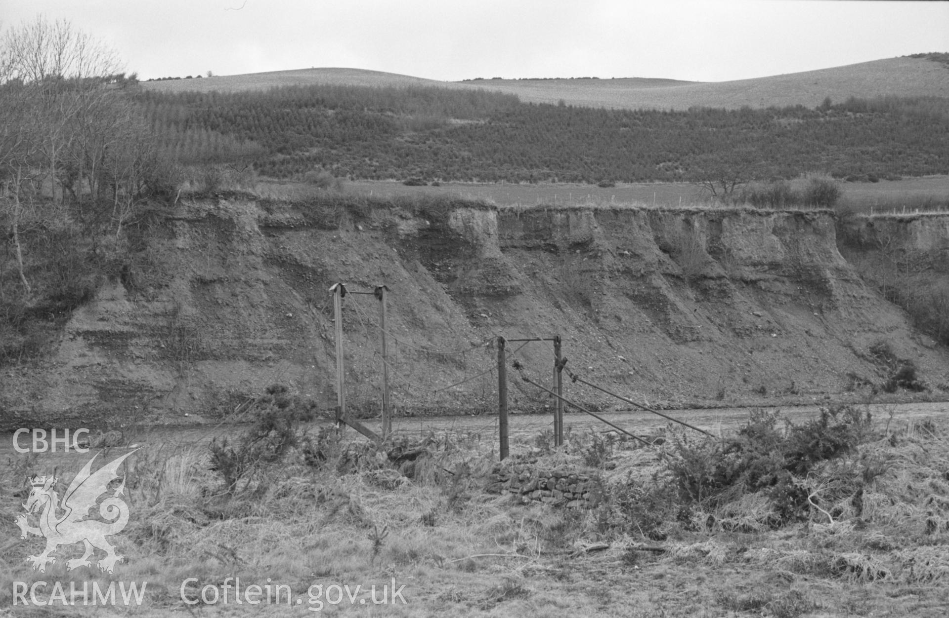 Digital copy of black & white negative showing remains of suspension footbridge across Ystwyth by the Halt. Coed Ty'n-Fron Llanddwy in distance on hillside. Photograph by Arthur Chater, 12th April 1965, from Grid Ref SN 6511 7445, looking north north east.