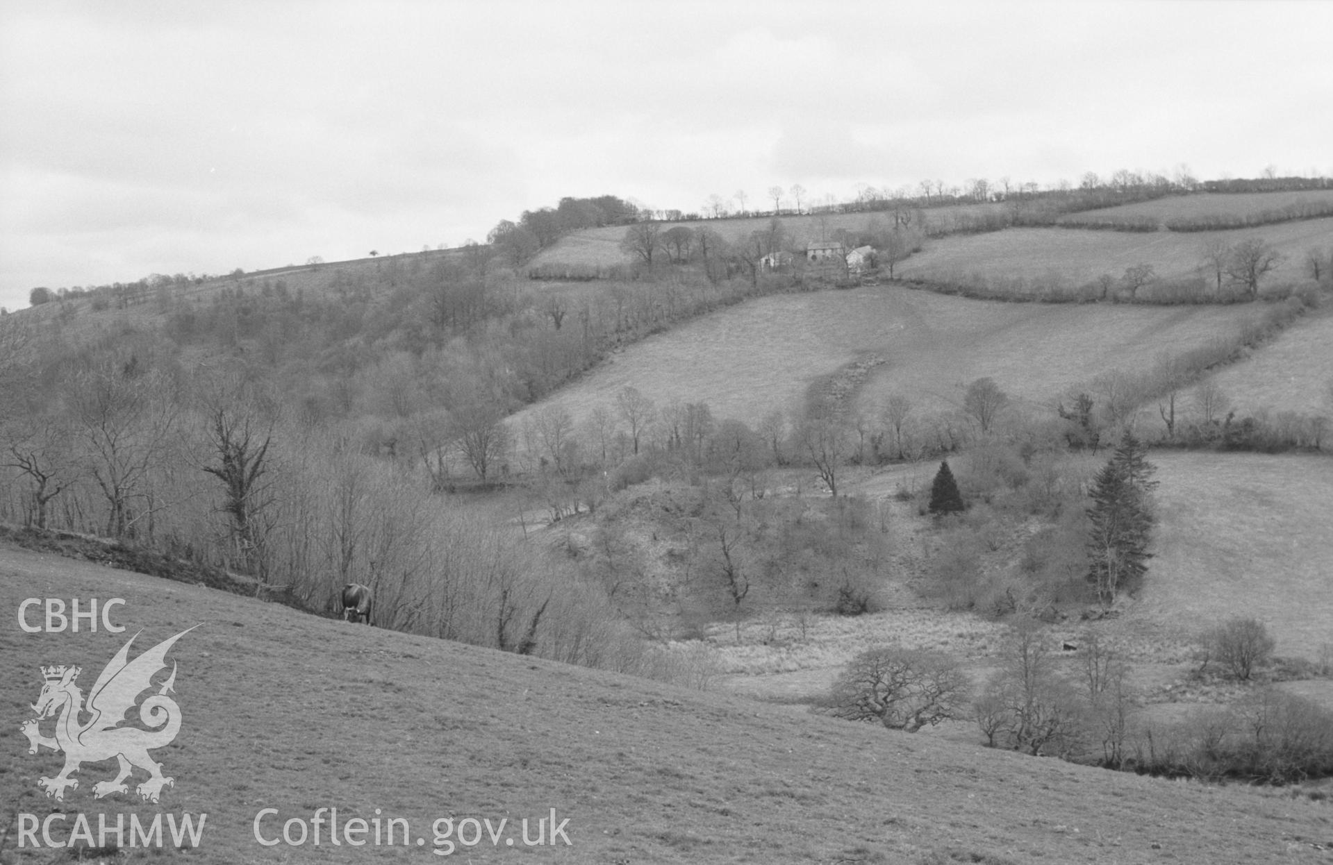 Digital copy of black & white negative showing side of Tomen Rhyd-Owen motte (in bottom of valley) with Brynclettwr farm above. Photographed by Arthur O. Chater in April 1965 from road near Llwynrhydowen (Grid Ref SN 4445 4400), looking south south west.