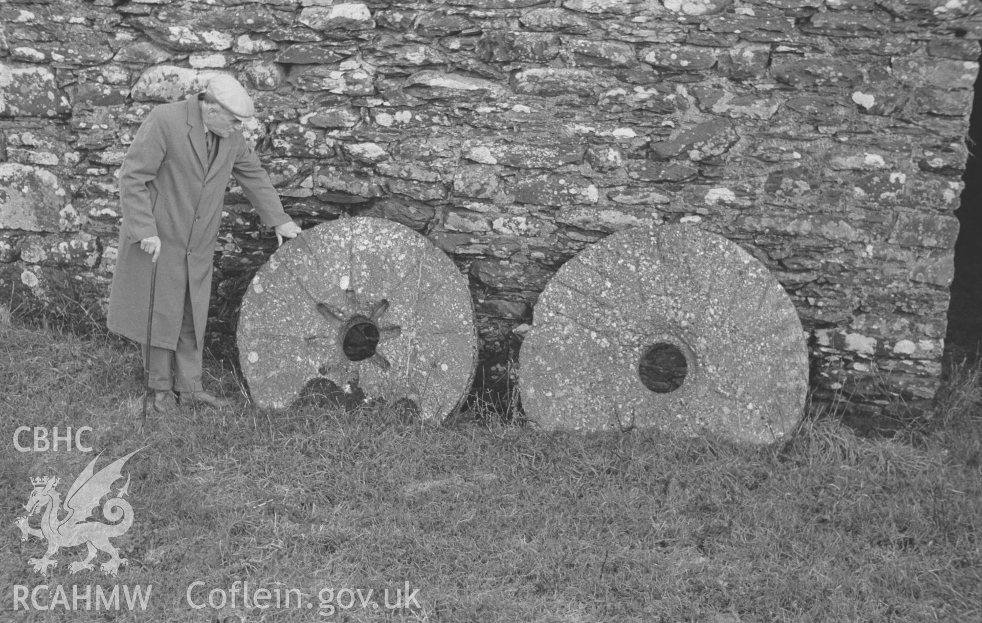 Digital copy of a black and white negative showing mill stones against the wall of the mill and a male figure at Swyddffynnon, north of Tregaron. Photographed in December 1963 by Arthur O. Chater from Grid Reference SN 6932 6614, looking south east.