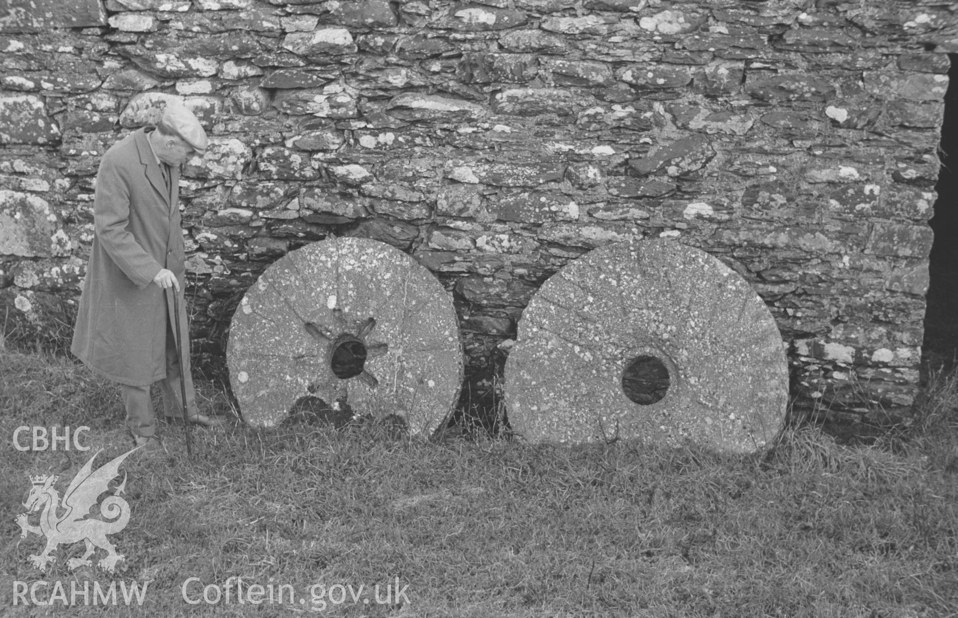 Digital copy of a black and white negative showing mill stones against the wall of the mill and a male figure at Swyddffynnon, north of Tregaron. Photographed in December 1963 by Arthur O. Chater from Grid Reference SN 6932 6614, looking south east.