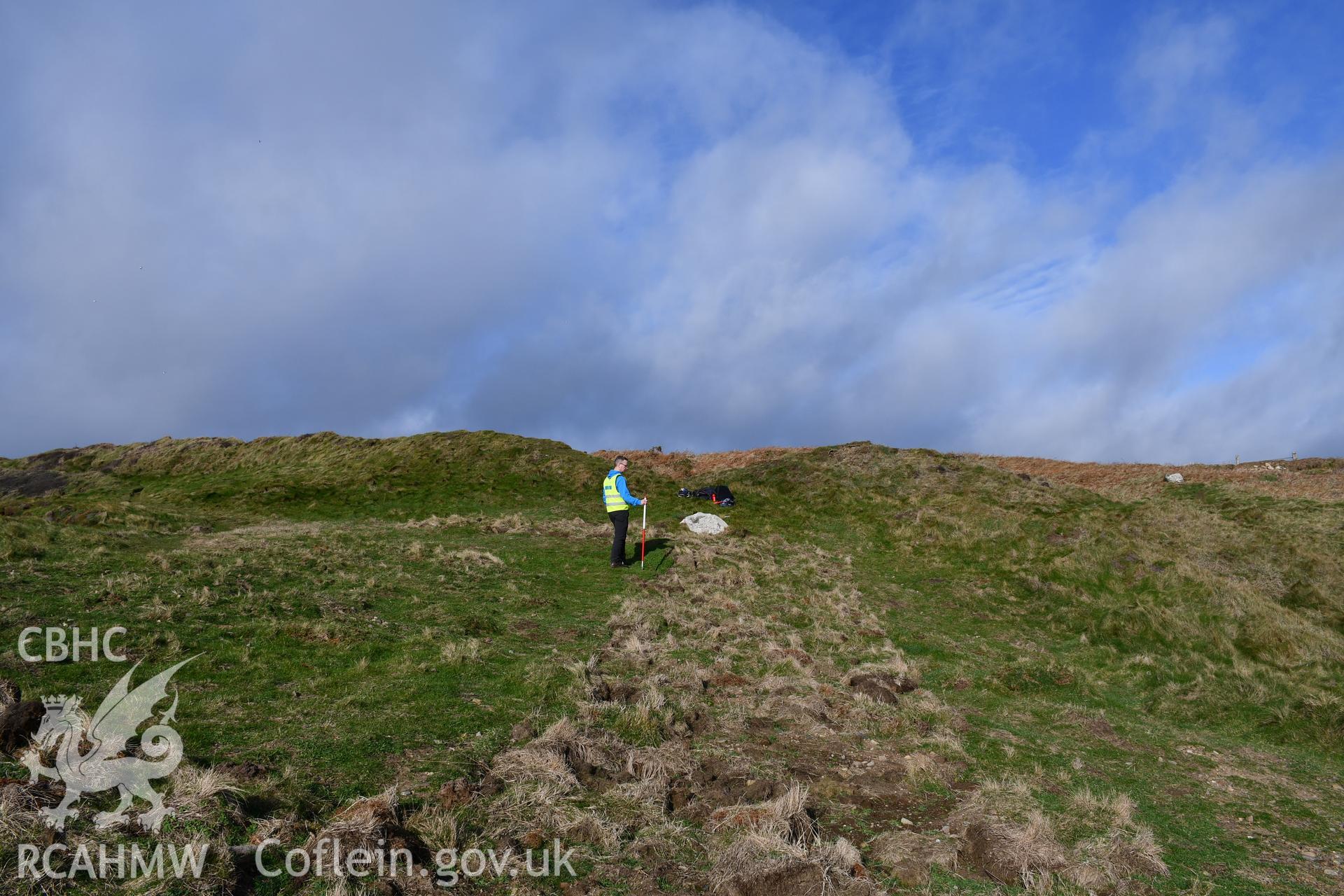 Reinstatement of turf over trench from Autumn 2021 excavation. Camera facing N. Person and 1m scale. Taken by Hannah Genders Boyd. Produced with EU funds through the Ireland Wales Co-operation Programme 2014-2023. All material made freely available throug