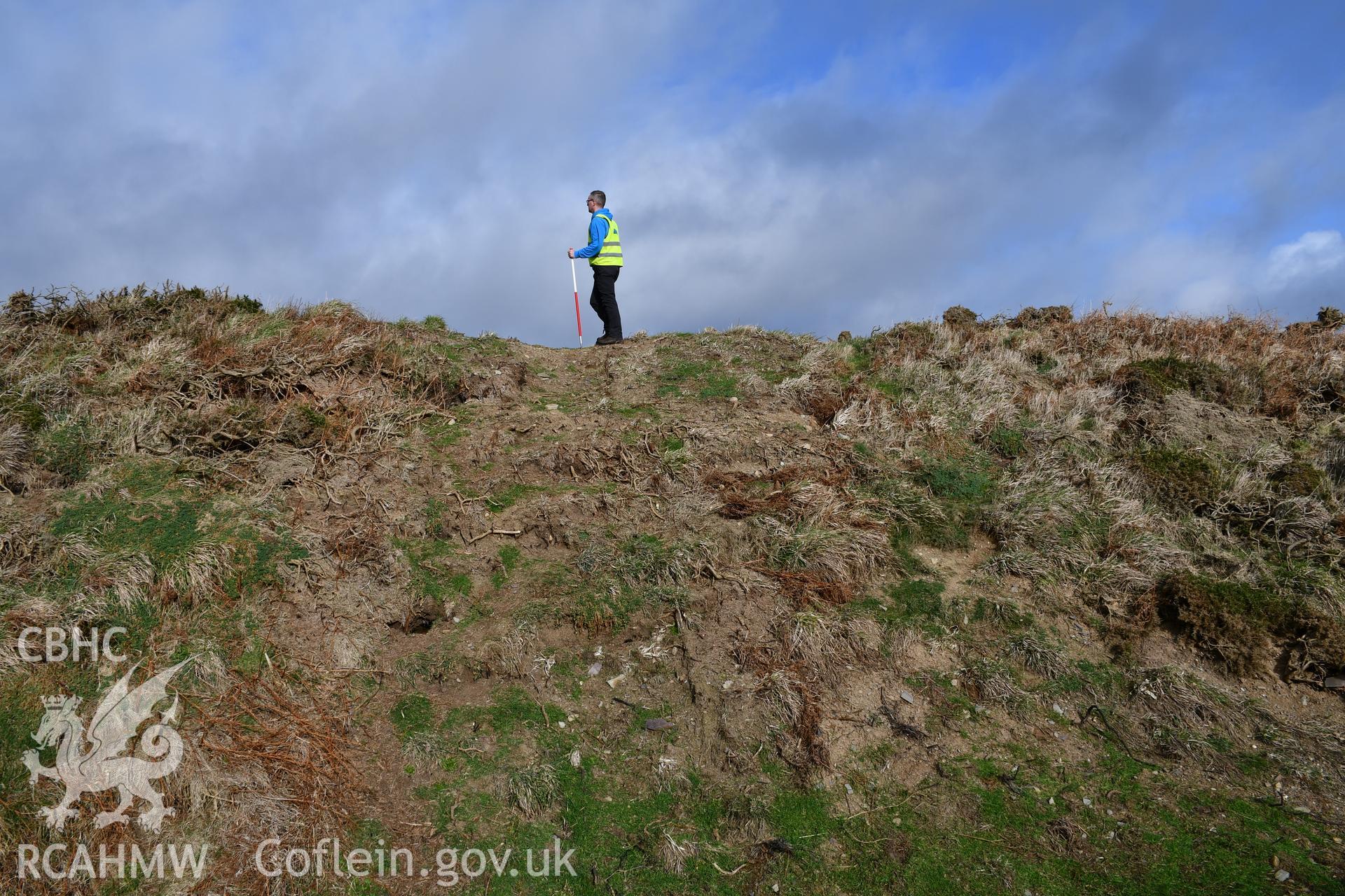 Reinstatement of turf over trench through rampart from Autumn 2021 excavation. Camera facing N. Person and 1m scale. Taken by Hannah Genders Boyd. Produced with EU funds through the Ireland Wales Co-operation Programme 2014-2023. All material made freely