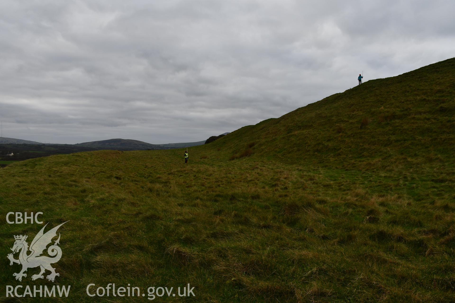 Outer ramparts and ditch of the hillfort, 2 figures and 2m scale. Taken by Hannah Genders Boyd. Produced with EU funds through the Ireland Wales Co-operation Programme 2014-2023. All material made freely available through the Open Government Licence.