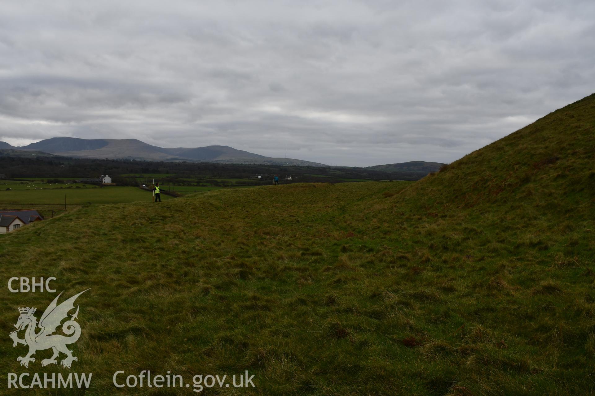 Outer ramparts and ditch of the hillfort, figure and 2m scale. Taken by Hannah Genders Boyd. Produced with EU funds through the Ireland Wales Co-operation Programme 2014-2023. All material made freely available through the Open Government Licence.