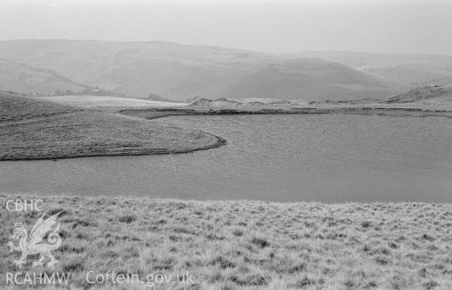 Digital copy of a black and white negative showing view looking across the Glog-Fawr mine reservoir. Ysbyty Ystwyth church on left (in distance); Maen Arthur wood beyond. Photographed by Arthur Chater on 14 April 1968. Looking north west from Grid Reference SN 750 708.