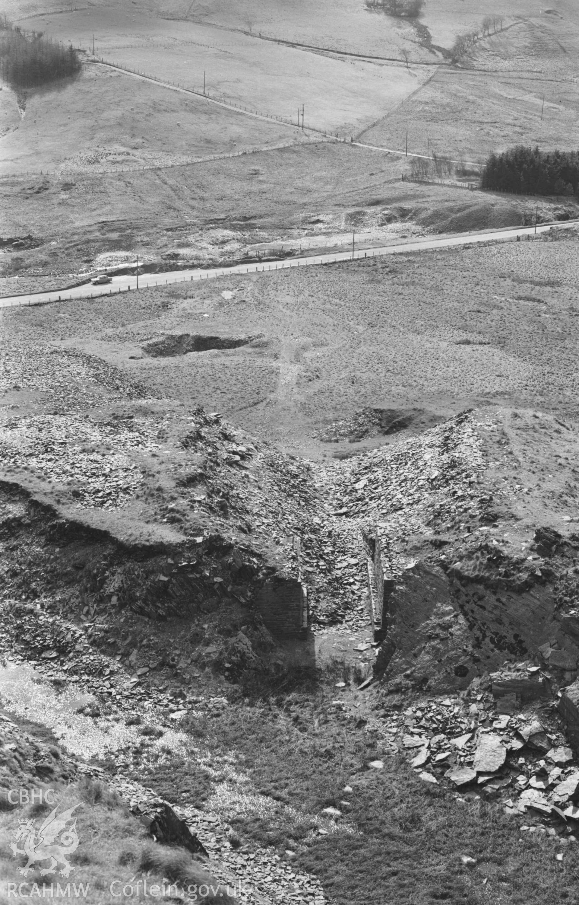 Digital copy of a black and white negative showing view across the entrance of the quarry behind Ffynnon-Cadno, showing the stone-lined entrance and the incline leading down the main road. Photographed by Arthur Chater on 2 April 1969. Looking south west from Grid Reference SN 7406 8087.