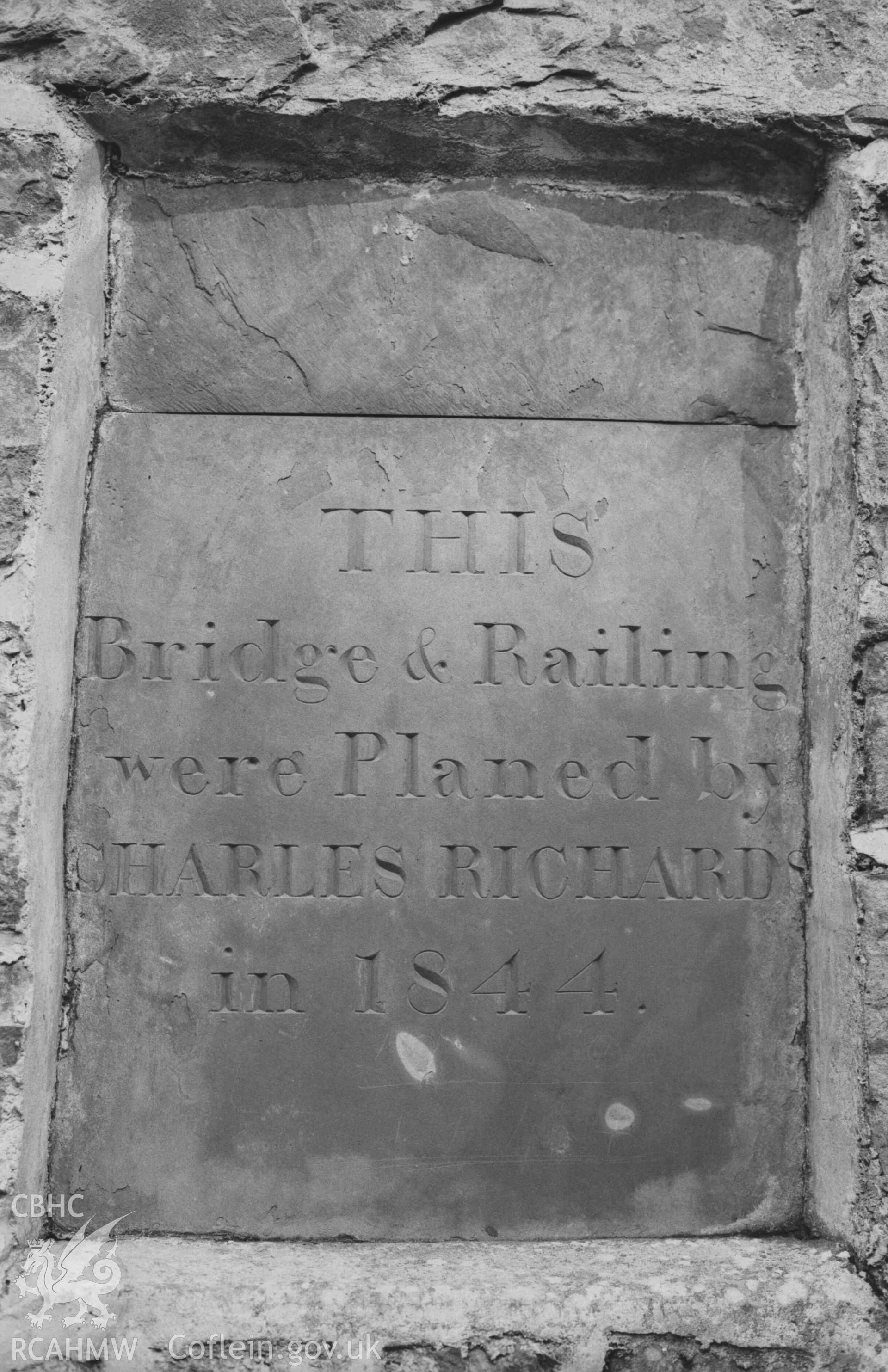 Digital copy of a black and white negative showing slate plaque on footbridge over the stream in Penglais Dingle, 200m south south west of Plas Penglais, at the south east corner of Dan-y-Coed housing estate. Photographed by Arthur Chater on 4 April 1969. Looking north east from Grid Reference SN 59340 8202. (Plaque reads: THIS Bridge & Railing were Planed by CHARLES RICHARD in 1844).